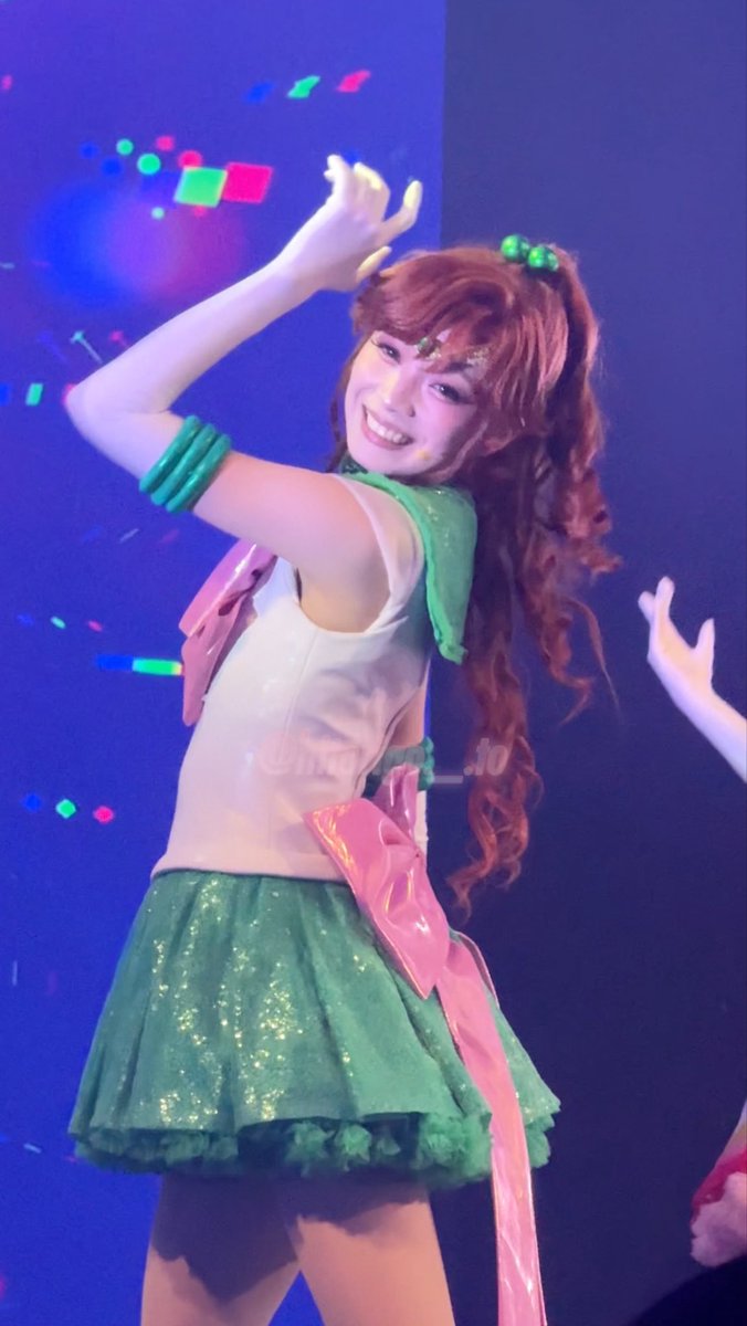 A cosplayer dressed as Sailor Jupiter from Sailor Moon poses with one arm raised, wearing the character's signature green skirt, white top, and pink bow on stage.