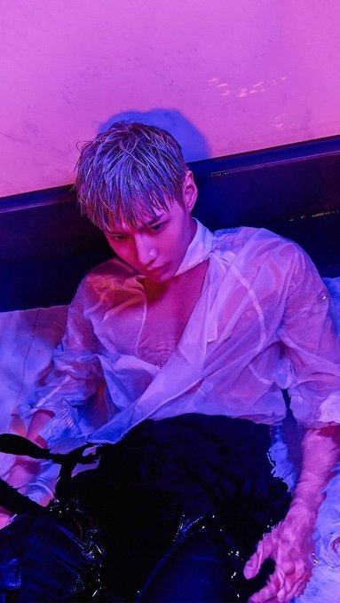 A person with wet hair wearing a partially open sheer white shirt sits in dim pink and purple lighting.