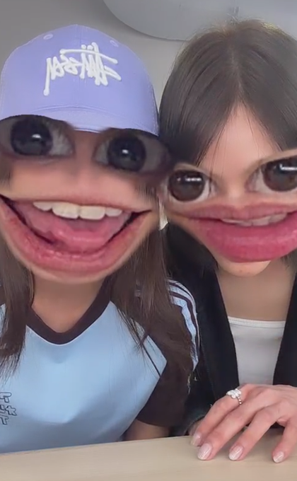 Two people wearing a face-distorting filter that enlarges their eyes and mouths, with one wearing a light purple Stussy cap and the other having dark hair and a ring on her finger.