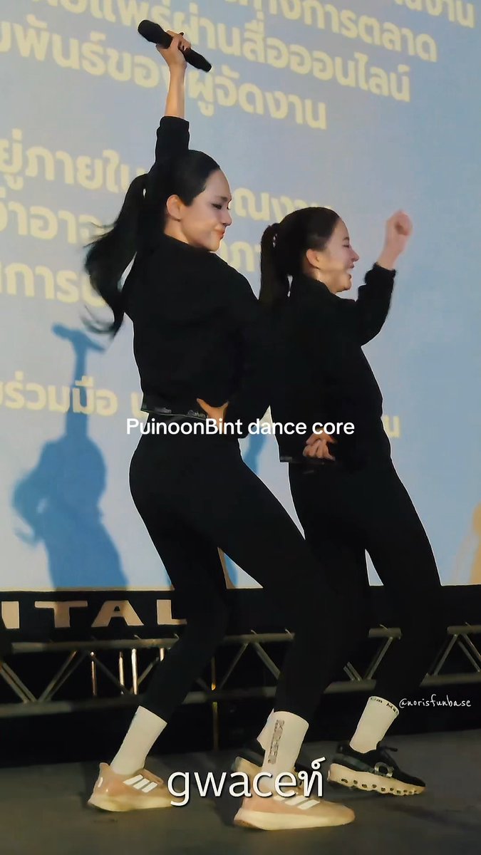 Two young women in matching black outfits with ponytails dancing back-to-back at an event, one holding a microphone; Thai text appears in the background, with overlays reading 'PuinoonBint dance core' and 'gwaceท์'.