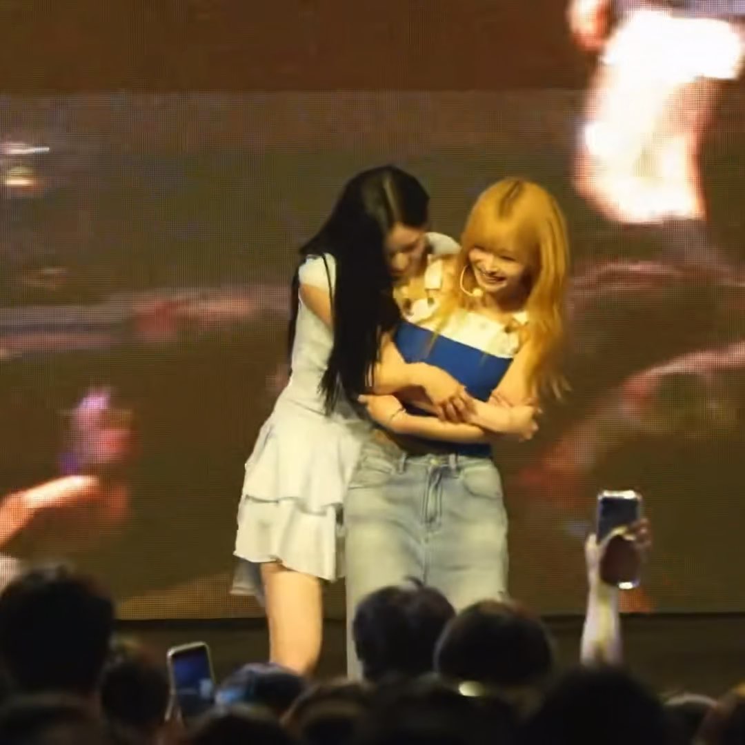 Two female performers share a warm hug on stage, one in a white ruffled dress with long dark hair and the other with long blonde hair wearing a blue top and denim jeans, with audience members holding up phones to capture the moment. The "Konnichiwa" caption suggests this is likely a K-pop concert greeting moment, reflecting the warm, friendly interaction between the two artists on stage.