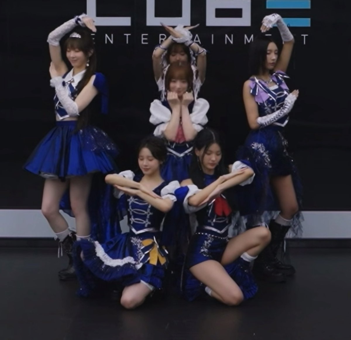 Five performers in matching navy blue and white costumes strike a choreographed pose on stage, with two kneeling in front and three standing behind, against a backdrop displaying partial "LBE ENTERTAINMENT" text. The image captures a moment from a LIGHTSUM performance, highlighting member Hina as mentioned in the post about the "City" (도시테) song's intro.