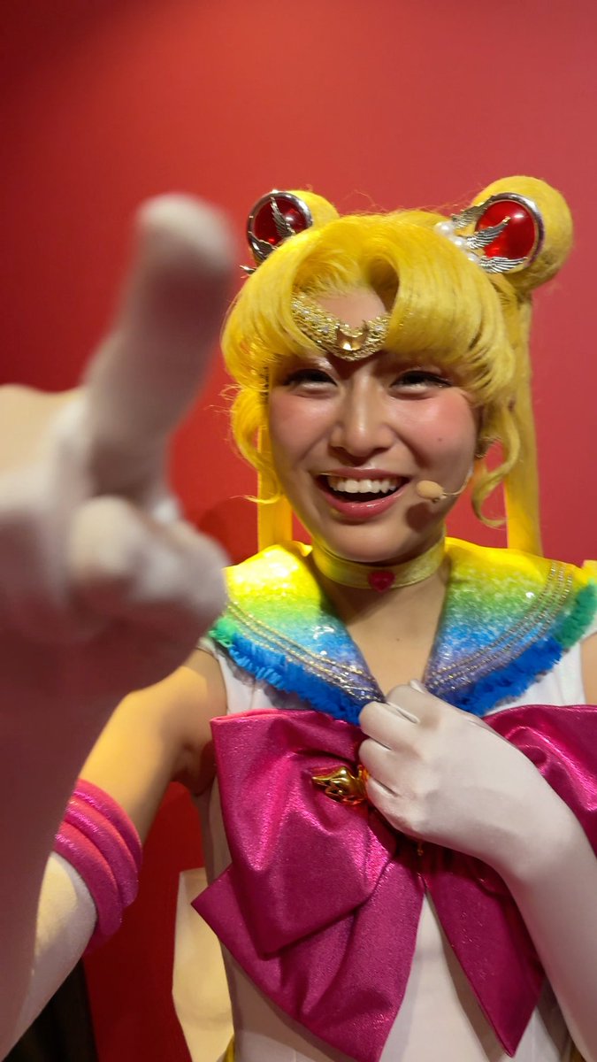 A woman in a Sailor Moon cosplay costume, complete with yellow odango buns, tiara, and white gloves, smiling and pointing toward the camera against a red background.