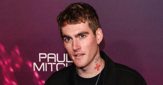A young man with short brown hair and facial tattoos poses in front of a purple backdrop with 'PAUL MITCHELL' branding.