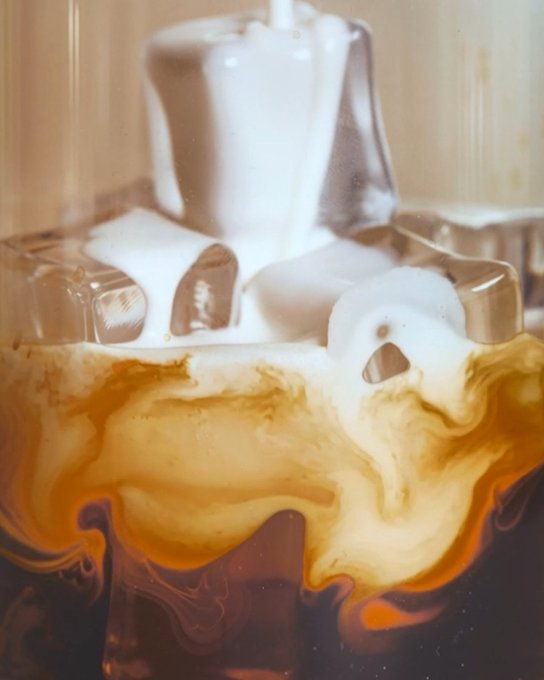 A close-up artistic shot of a beverage showing ice cubes with white cream and a brown, caramel-colored liquid swirling together beneath the surface.