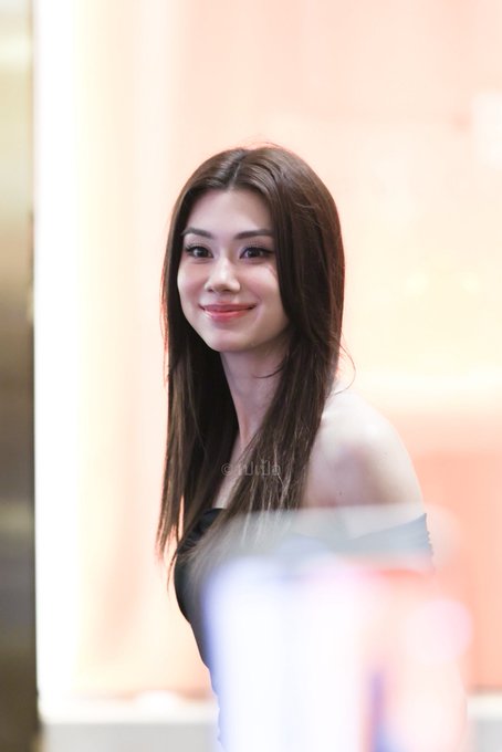 A smiling young woman with long brown hair poses for the camera, wearing a strapless top against a blurred background with a faint watermark.