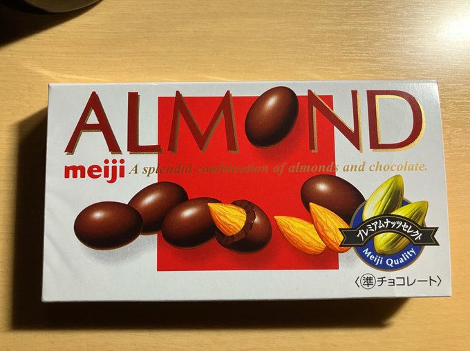 A Meiji Almond Chocolate box rests on a wooden surface, featuring the product name "ALMOND" prominently along with images of chocolate-covered almonds and a "Meiji Quality" premium seal. The "Johnny Joestar" reference suggests a fan sharing this Japanese chocolate treat, possibly as a nod to the *JoJo's Bizarre Adventure* character's connection to the brand name.
