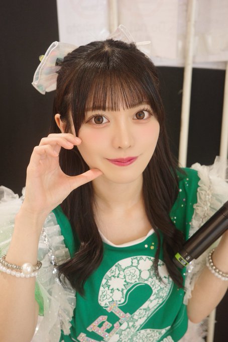 A close-up portrait of a young woman with dark hair and bangs wearing a green outfit with a bow, posing with her hand near her face.