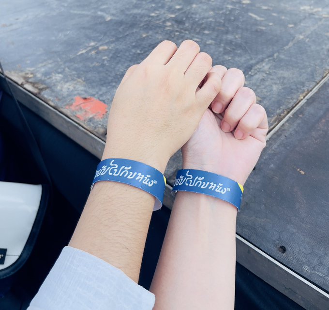 A close-up photo of two hands held together, both wearing matching blue wristbands with white Thai script.