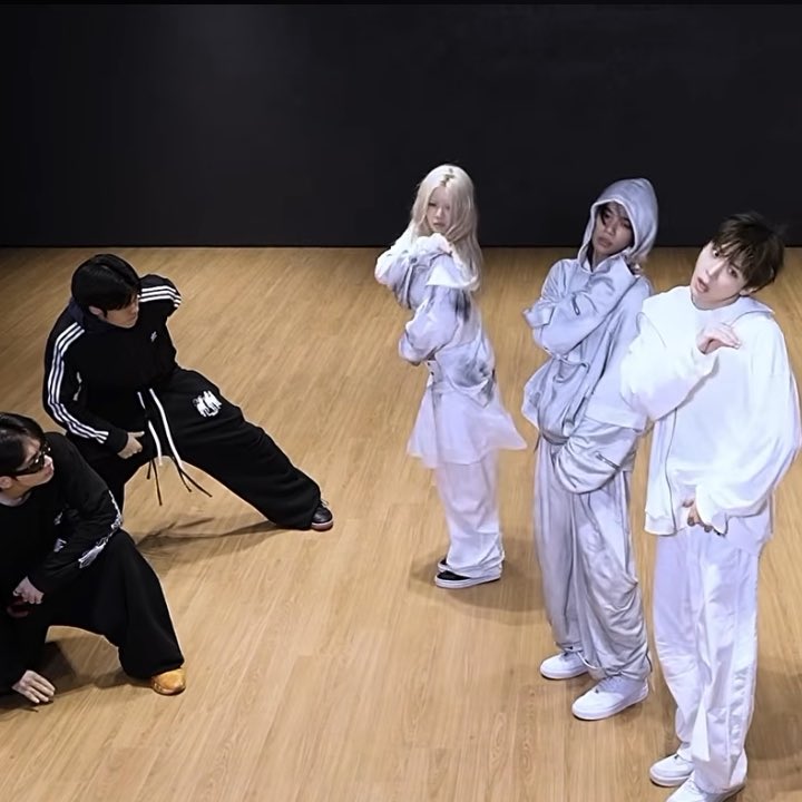 A K-pop dance practice room scene with dancers in black tracksuits and others in white hoodies posing on a wooden floor against a black backdrop.