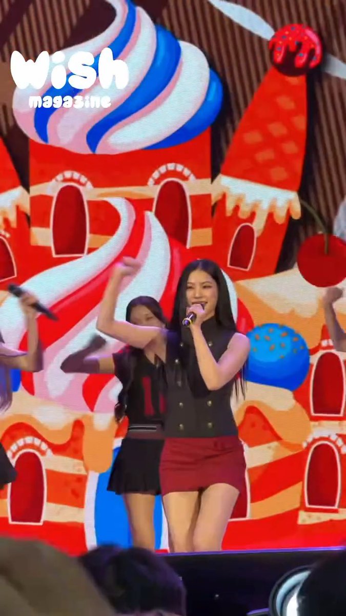 A female performer sings into a microphone while dancing on stage in a black top and red skirt, with backup dancers and a colorful candy-themed backdrop; a 'Wish magazine' logo is in the corner.