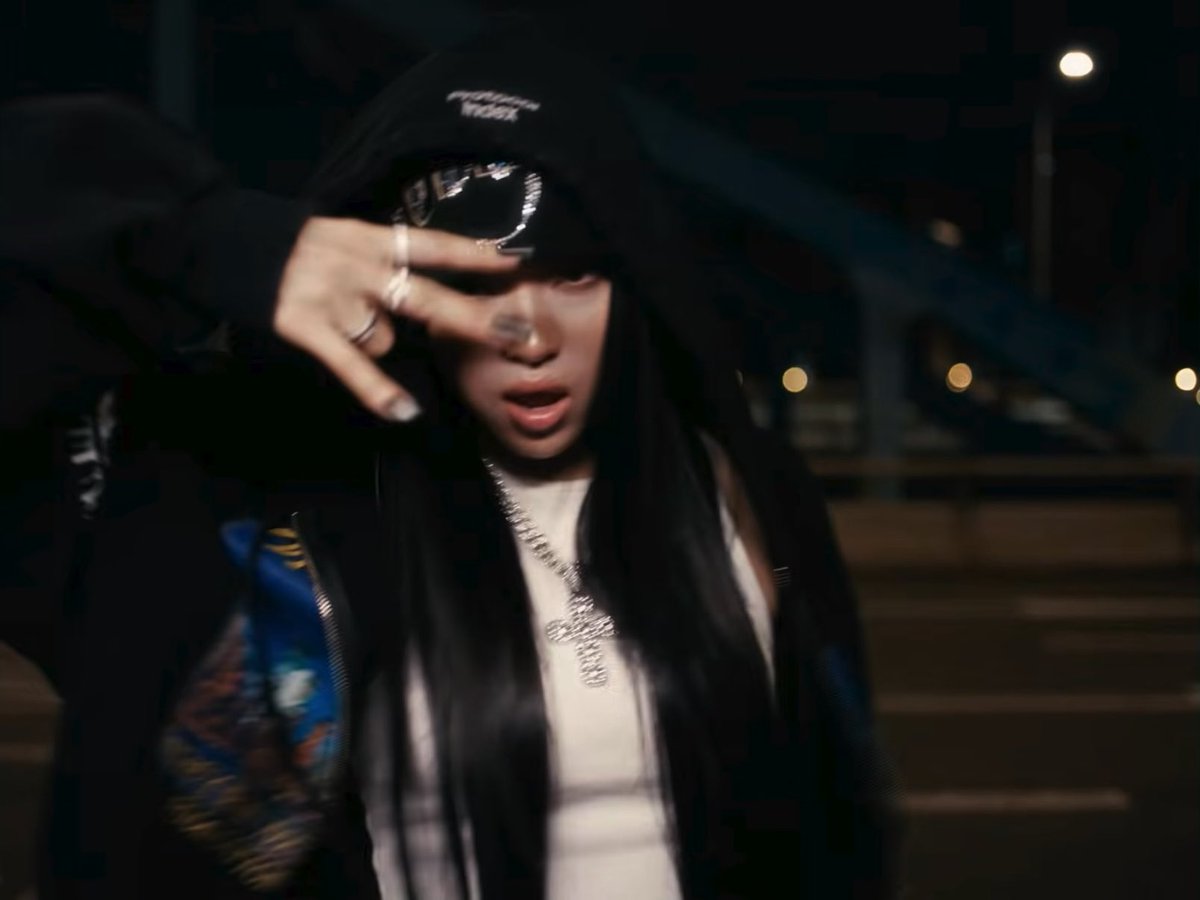 A young woman wearing a black cap and an icy diamond-encrusted pendant makes a peace sign in a dimly lit urban nighttime setting, with streetlights blurred in the background. The post's reference to "dogs" likely plays on hip-hop slang, where the rapper (appearing to be GloRilla) is showcasing her confident, glamorous style and commanding presence.