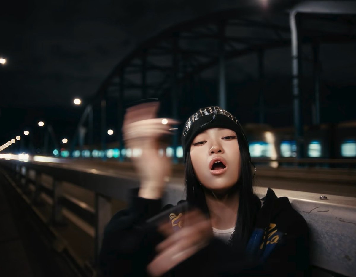 A woman wearing a black Christian Dior beanie and hoodie takes a selfie at night on a bridge, with city lights and a bridge structure visible in the background.