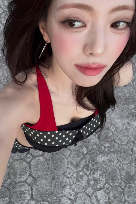 A close-up selfie of a woman with pink blush and eyeliner, wearing a black and white polka dot top with a red accent.