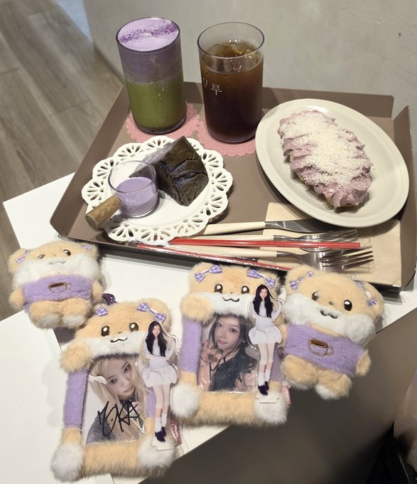 A table spread featuring purple-themed drinks, desserts, and cute plushies in orange and purple colors alongside acrylic photocards of K-pop idols.