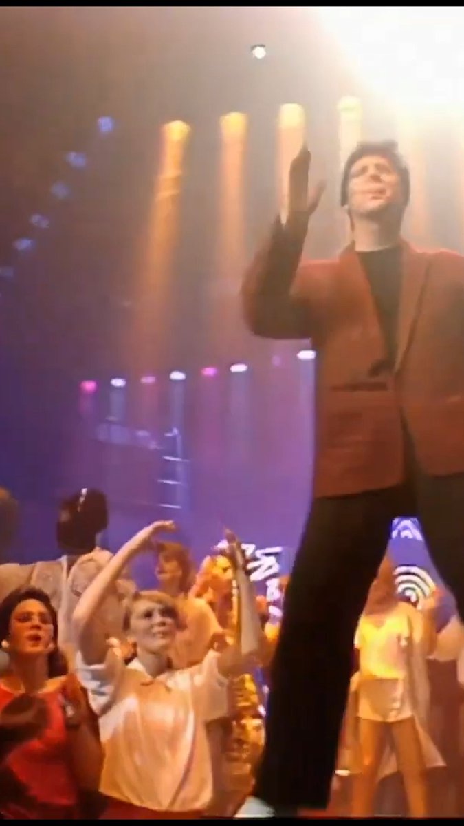 A performer in a rust-colored blazer and dark trousers waves to an enthusiastic crowd on a vibrant stage lit with orange spotlights, with a BBC logo visible on a backdrop. The scene captures a classic televised performance setting reminiscent of 1980s UK music shows, fitting the era when Shaky's cover of "Lipstick, Powder and Paint" climbed to No. 14 on the UK Singles Chart.