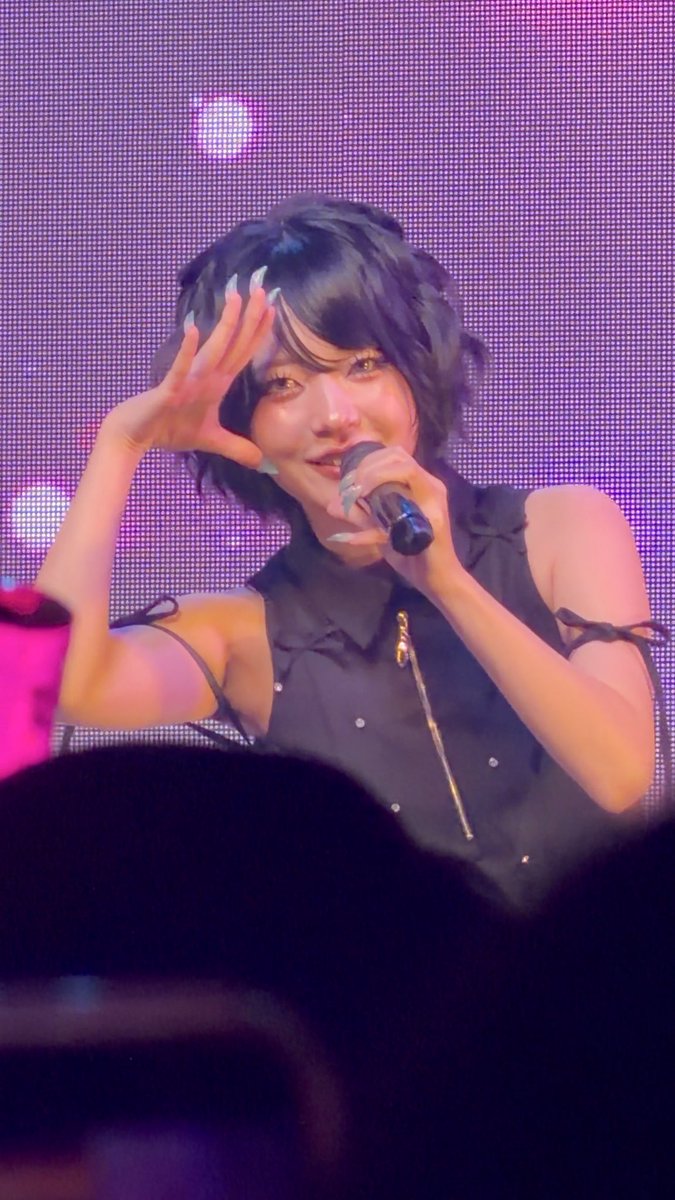 A performer with short dark hair sings into a microphone while making a hand gesture, captured in a close-up shot on a purple-lit stage. The caption's note about "約9倍" (approximately 9x zoom) explains the tight framing that makes the subject's face and manicured nails so detailed, prompting the "brain-chemical rush" reaction from the photographer.
