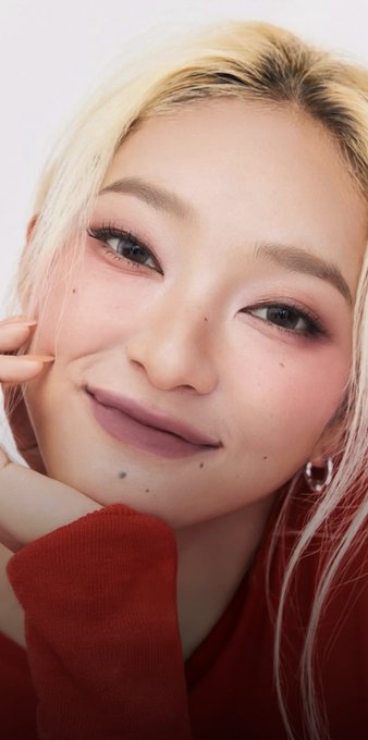A close-up portrait of a smiling young woman with blonde hair, wearing a red long-sleeved top, pink eyeshadow, and mauve lipstick.