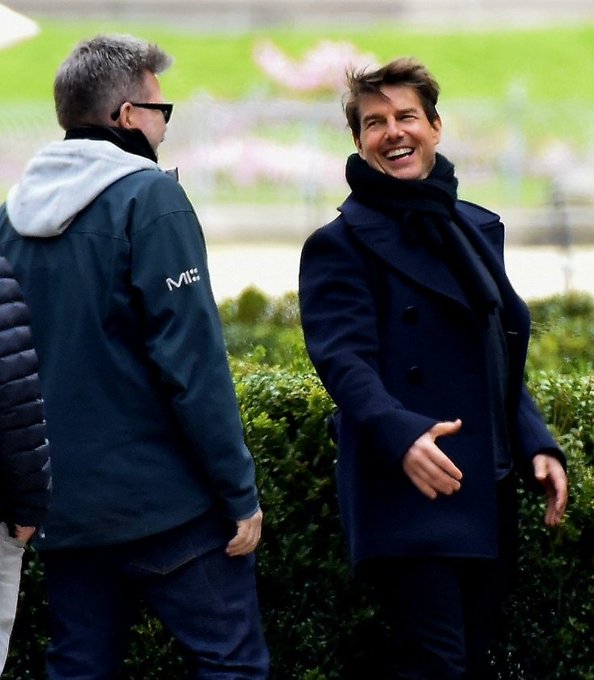 Actor Tom Cruise, wearing a dark coat and scarf, laughs alongside crew members outdoors near hedges.