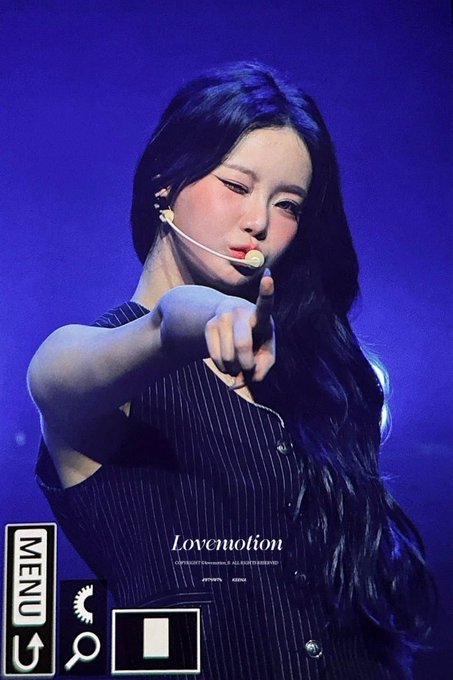 A female performer with long dark hair and a headset microphone points directly at the camera while wearing a pinstriped sleeveless top, with a 'MENU' graphic and 'Lovemotion' watermark visible on the image.