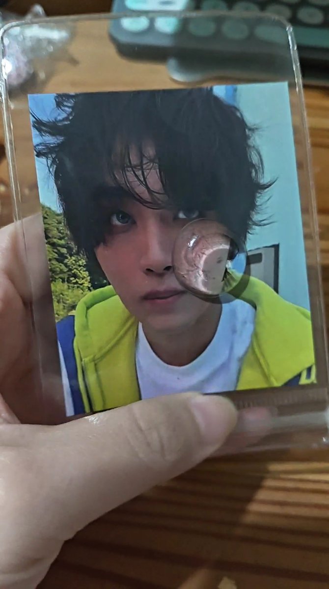 A hand holds a photocard of a young man with dark hair wearing a yellow hoodie and white shirt.