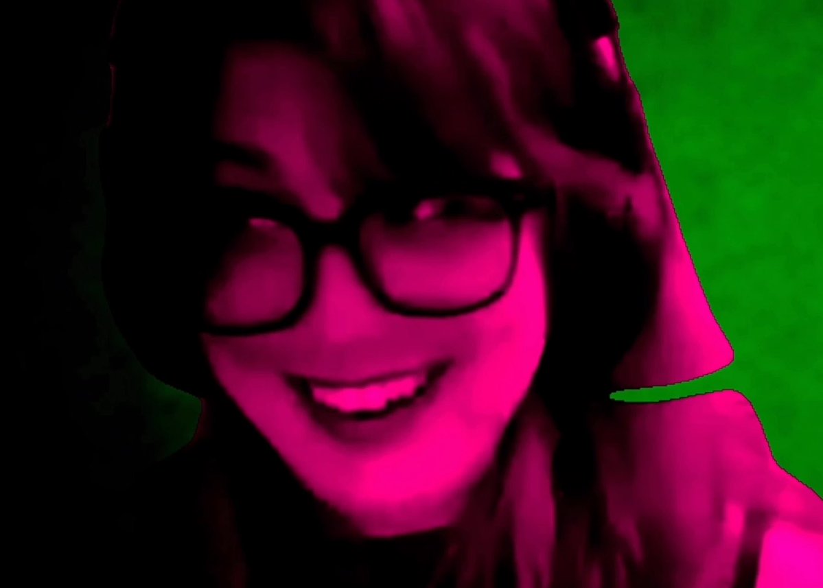 A person with dark hair and glasses smiling, bathed in heavy pink and green lighting.
