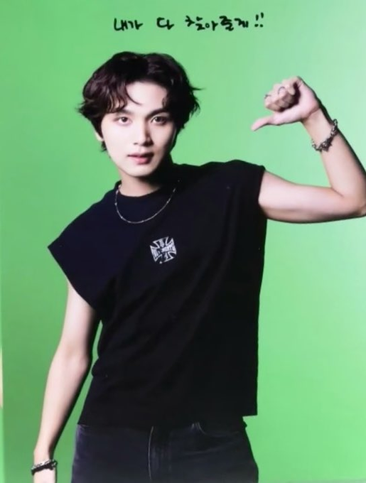 A young man with dark wavy hair poses against a green screen, flexing his arm while wearing a black sleeveless shirt with a British flag logo; Korean text at the top reads "내가 더 잘어울려!!" (It suits me better!!).
