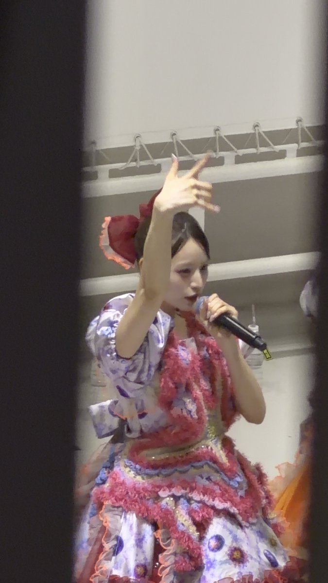 A performer in an elaborate purple and pink ruffled dress with a large red bow in her hair, holding a microphone and gesturing while on stage.