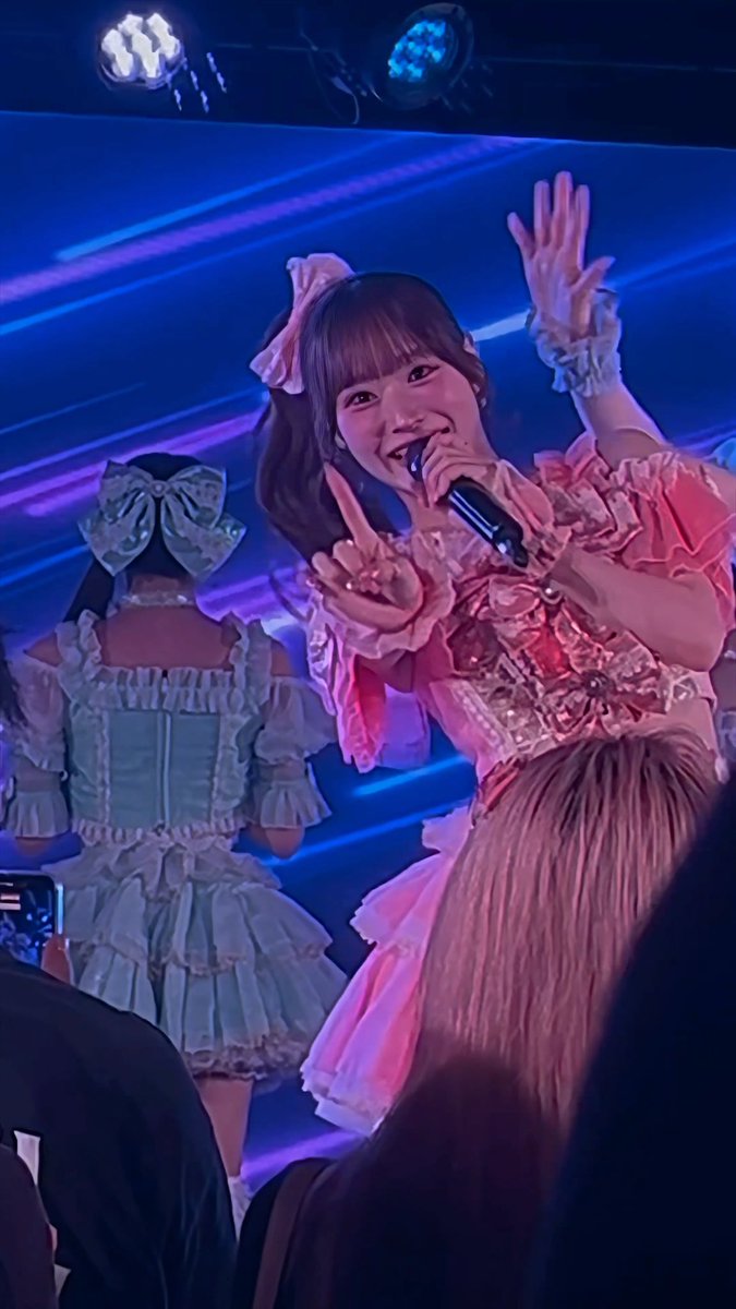 A young woman in a frilly pink outfit and bow poses on stage with her hands raised while singing into a microphone, alongside another performer in a blue outfit.