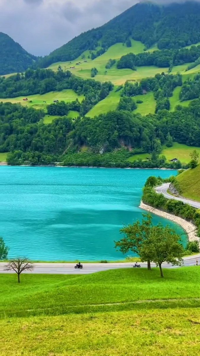 Two riders cruise along a winding alpine road beside a striking turquoise lake, surrounded by lush green hillsides and forested mountains — a scene that looks like it could be straight out of Switzerland. The vibrant landscape perfectly matches the post's celebration of stunning nature and travel.