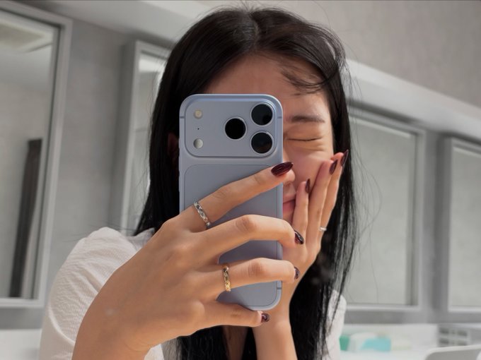 A young woman with long dark hair takes a mirror selfie with a lavender smartphone, covering her face while displaying dark red nail polish and two rings on her fingers.