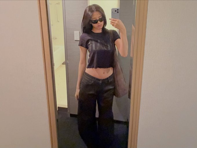 A mirror selfie of a stylish young woman wearing sunglasses, a dark cropped t-shirt, and baggy black jeans, carrying a brown shoulder bag.
