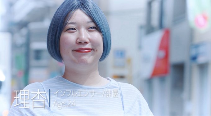 A smiling young woman with blue hair, identified as "Risa / Influenza Fujou" (Age: 24), stands in an urban street setting. The text overlay and her cheerful demeanor suggest she is one of the participants or talents featured in this homemade "Renai Reality" production.