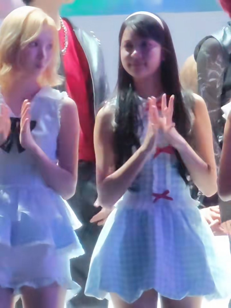 Two young girls in matching white dresses with red bow accents stand side by side, clapping, with the girl on the right appearing to glance sideways while her companion stares straight ahead with a blank expression—matching the post's playful commentary about Jiu sneaking peeks at a seemingly oblivious Lemen.