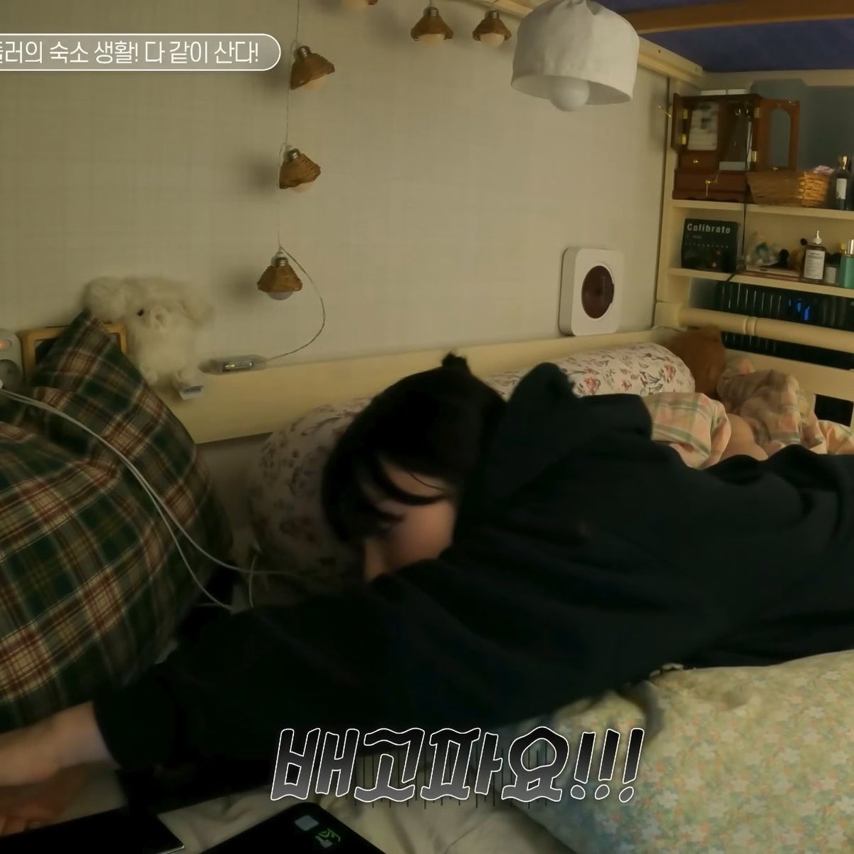 A person with dark hair in a black hoodie lies face-down on a bed in a room with string lights, while Korean text reads '리라의 숙소 생활! 다 같이 산다!' at the top and '배고파요!!!' at the bottom.