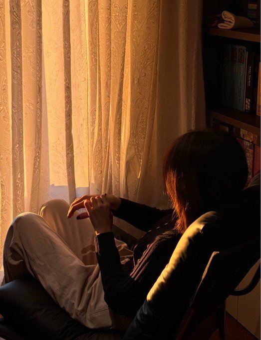 A silhouette of a woman sitting in a chair by a window with sheer curtains, bathed in warm golden sunset light, with a bookshelf visible to the right.