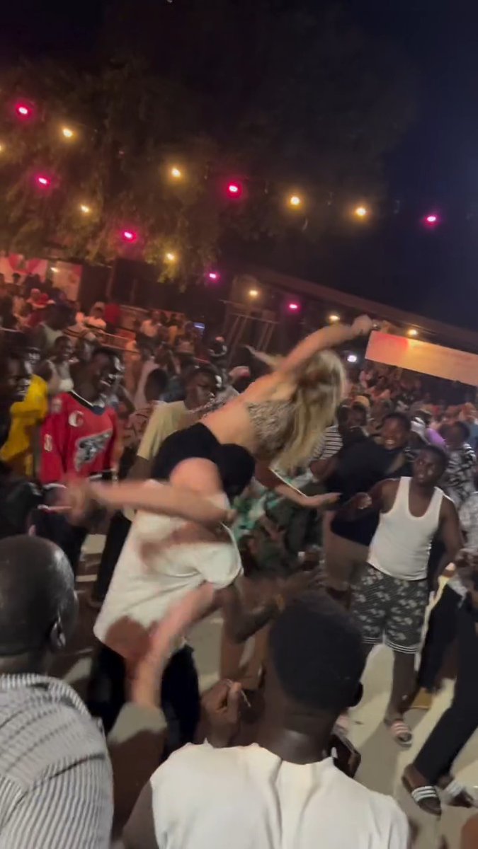A nighttime outdoor scene of a crowded party or concert with string lights overhead and a blonde woman being lifted or dancing above the crowd.