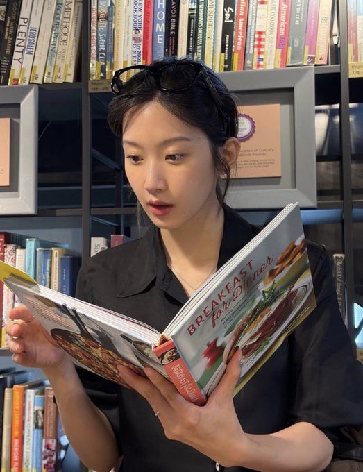 A young woman with sunglasses perched on her head reads a cookbook titled "BREAKFAST for Dinner" in what appears to be a library or bookstore setting with bookshelves behind her.
