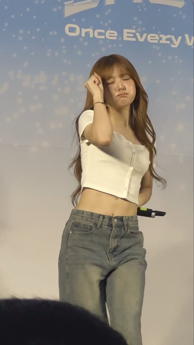 A young woman with long brown hair wearing a white crop top and blue jeans, appearing emotional with closed eyes and hand near her face, photographed against a backdrop reading 'Once Every'.