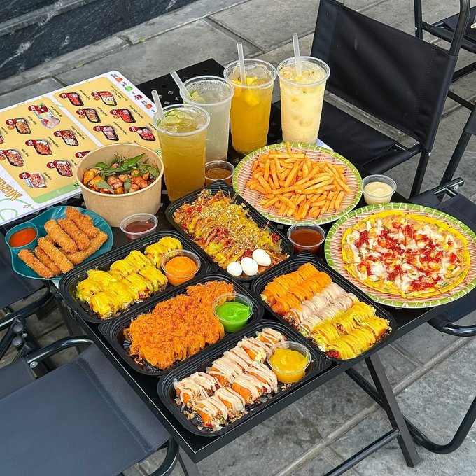 An outdoor table filled with takeout containers of sushi, rice bowls, french fries, fried snacks, and iced beverages, accompanied by a menu.