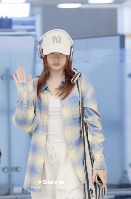 A stylish young woman wearing a white "NY" baseball cap and headphones poses with a peace sign, dressed in a yellow and blue plaid flannel shirt (THISISNEVERTHAT's Flannel Check Shirt) over a white top, complementing the featured product link in the post. The image showcases the casual Korean streetwear aesthetic, with a watermark "Salvia" visible at the bottom.
