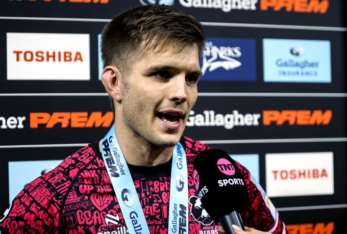 A Bristol Bears rugby player, wearing a pink and black patterned team shirt with a Gallagher Premiership lanyard, is being interviewed by TNT Sports with a microphone in front of a sponsor backdrop featuring Toshiba and Gallagher logos. The image illustrates the club's new NFL-style media access initiative, showing players like this one being made available to journalists for post-match interviews in a designated mixed zone near the changing room at Ashton Gate.