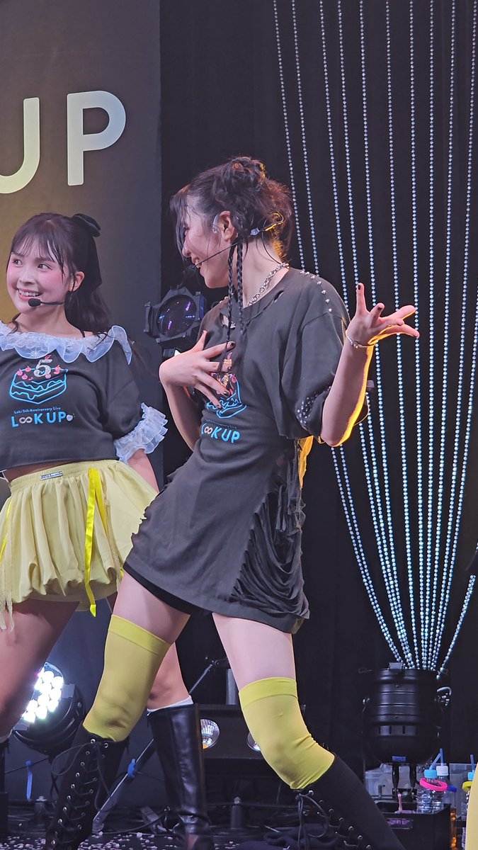 Two female performers on stage wearing matching dark shirts with a birthday cake logo and 'LOOK UP' text, paired with yellow skirts and knee-high socks, performing at a concert event.