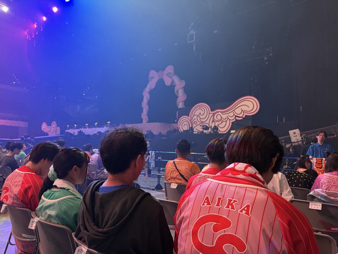 Audience members wearing colorful jerseys sit facing a stage with elaborate swirling decorative elements lit by purple stage lights.