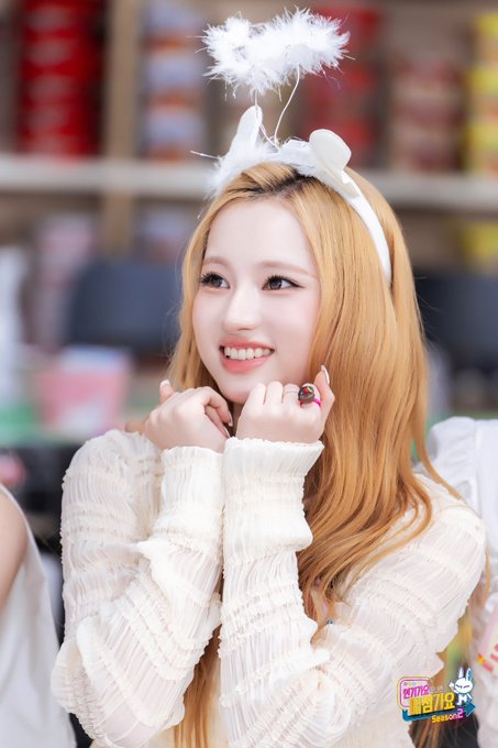 A smiling young woman with long blonde hair wears a white headband with fluffy white decorations and a cream-colored sweater, with 'Music Bank' branding visible at the bottom right.