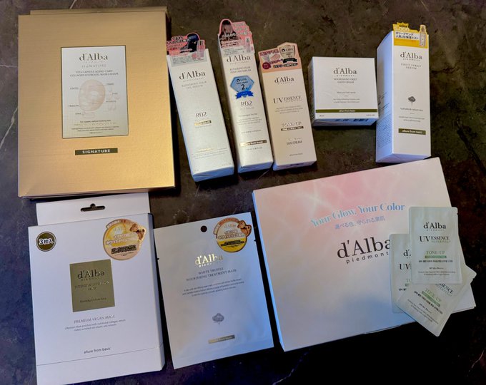 An assortment of d'Alba skincare and beauty products, including the signature White Truffle collection, various sheet masks, and toner-up ampoules, are laid out on a dark surface.