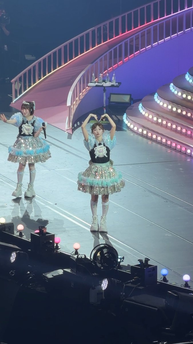 Two female performers in sparkly turquoise and white outfits perform on a stage with a pink-lit staircase in the background.