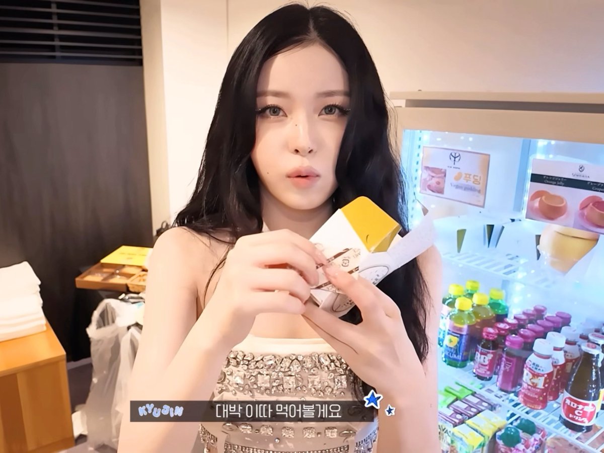 A young woman with long dark hair in a sparkly top holds a yellow box of snacks, with Korean text overlay '대박 이따 먹어볼게요' and 'NYLON' visible, with a refrigerated display of drinks behind her.
