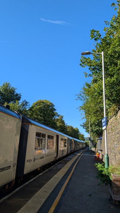 A train is stopped at a station platform under a clear blue sky, with a tall street lamp, green trees, and a stone wall visible alongside the tracks.