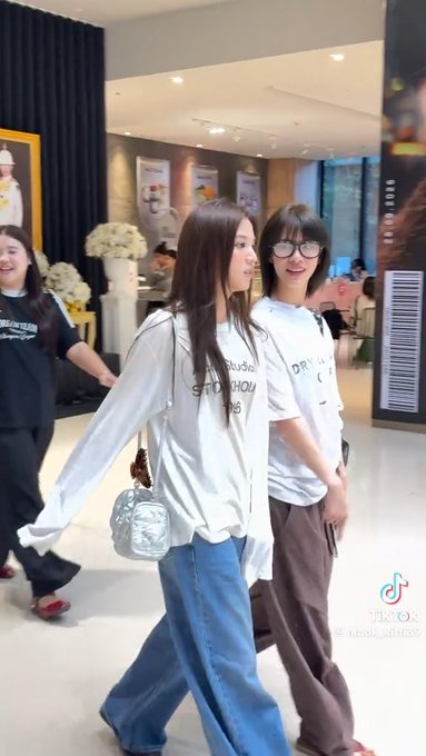 Two young people walk together in a brightly lit indoor shopping or hotel area; the person on the left wears a white long-sleeve graphic shirt, while the person on the right wears sunglasses and a white t-shirt with brown pants.